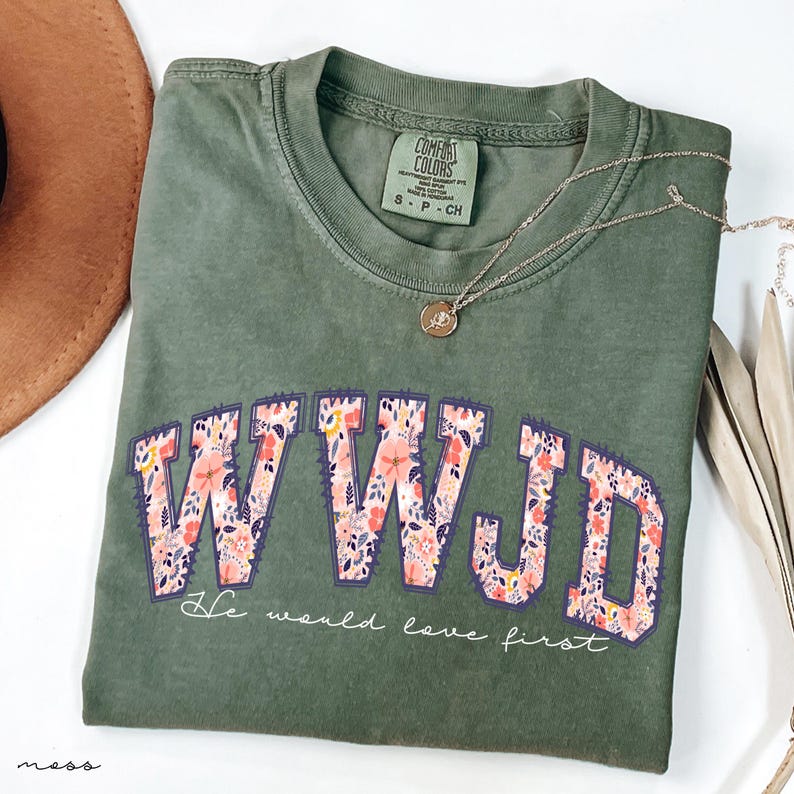 May include: An olive green t-shirt featuring the letters WWJD in a floral design. The text "He would love first" is below the letters. A silver necklace with a pendant is on the shirt.