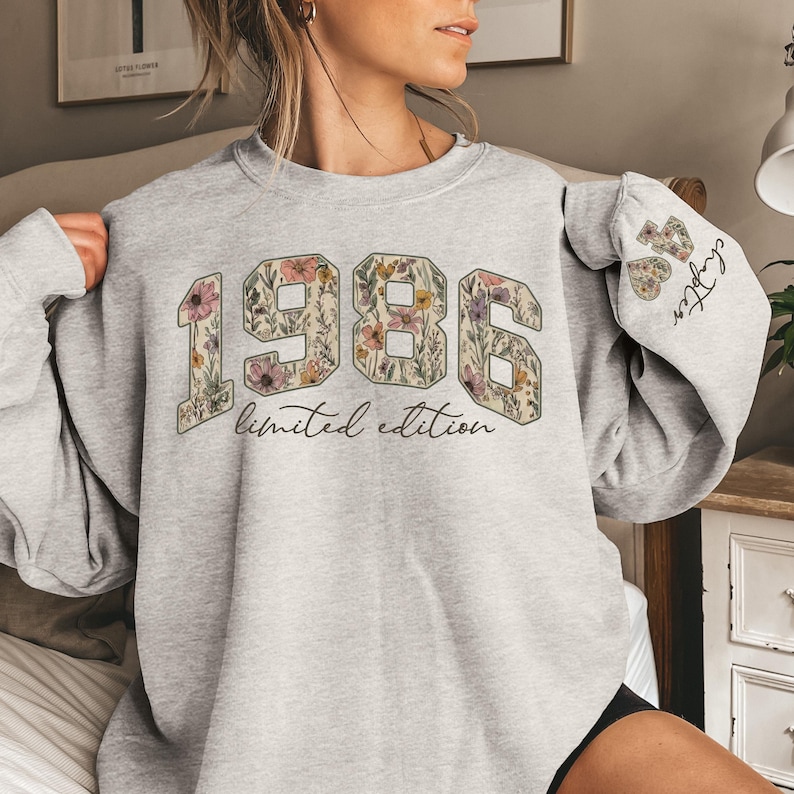 Puede incluir: Una sudadera gris claro con el a&ntilde;o "1986" en una gran impresi&oacute;n floral. Las palabras "limited edition" est&aacute;n debajo de los n&uacute;meros. La manga tiene las palabras "48 chapter" en un dise&ntilde;o floral.