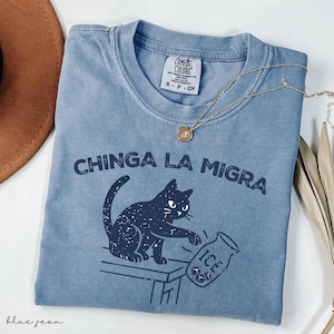 May include: A blue jean Comfort Colors t-shirt with the phrase "CHINGA LA MIGRA" and a black cat graphic. The cat is knocking over a jar of ice. A gold necklace and a brown hat are also visible.
