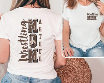 Puede incluir: Camiseta blanca con texto marrón que dice "Wrestling Mom" en la parte delantera y "Wrestling Mom" con un gráfico de un luchador en la parte trasera.