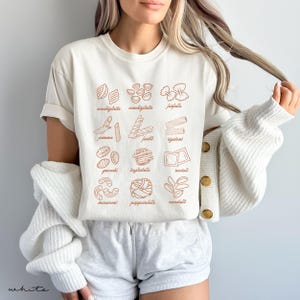 Op de afbeelding: Een wit T-shirt met een afbeelding van verschillende soorten pasta, elk met de naam in bruine tekst. De pastasoorten zijn:  conchiglie, farfalle, pennes, fusilli, rigatoni, gnocchi, tagliatelle, ravioli,  macaroni, pappardelle en cavatelli.