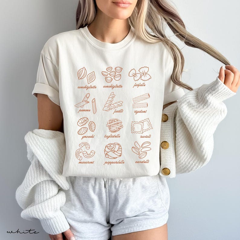 Op de afbeelding: Een wit T-shirt met een afbeelding van verschillende soorten pasta, elk met de naam in bruine tekst. De pastasoorten zijn:  conchiglie, farfalle, pennes, fusilli, rigatoni, gnocchi, tagliatelle, ravioli,  macaroni, pappardelle en cavatelli.