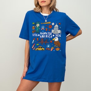 K&ouml;nnte beinhalten: Ein k&ouml;nigsblaues T-Shirt mit wei&szlig;em Rundhalsausschnitt. Es zeigt ein farbenfrohes Grafikdesign mit dem Text "Happy 250 America 2026" und Illustrationen der Freiheitsstatue, eines Adlers und anderer patriotischer Symbole.