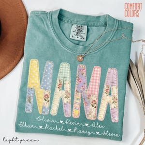 May include: Light green Comfort Colors t-shirt with the word "NANA" in patchwork letters. The letters are decorated with floral and patterned fabrics. Below the word are names and hearts. A silver necklace is draped across the shirt.