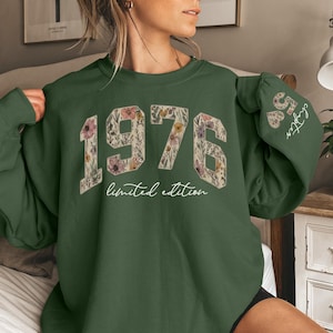 May include: A dark green sweatshirt with the year "1976" in large, floral-patterned numbers. The words "limited edition" are below the numbers. The sleeve has the text "58 Chapter" in white. The sweatshirt is a casual style.