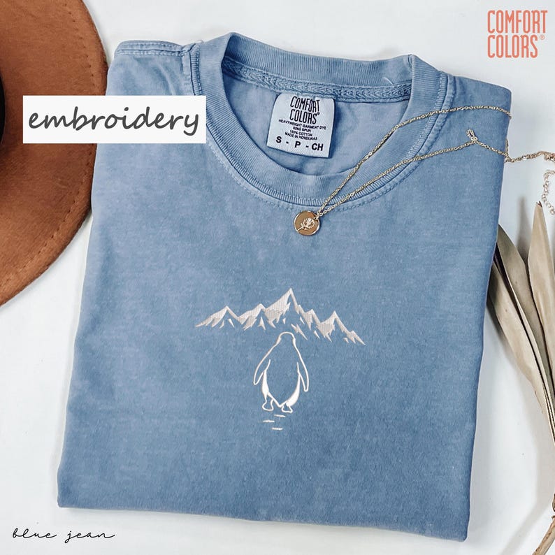 May include: Blue jean Comfort Colors t-shirt with an embroidered penguin and mountain design. A gold necklace with a pendant is draped across the shirt. The word "embroidery" is written on a white rectangle.