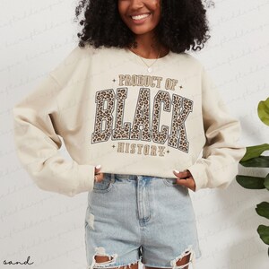 May include: Sand-colored sweatshirt with the text "PRODUCT OF BLACK HISTORY" in a bold, leopard print design. The sweatshirt has long sleeves and a crew neck. The overall style is casual and comfortable.