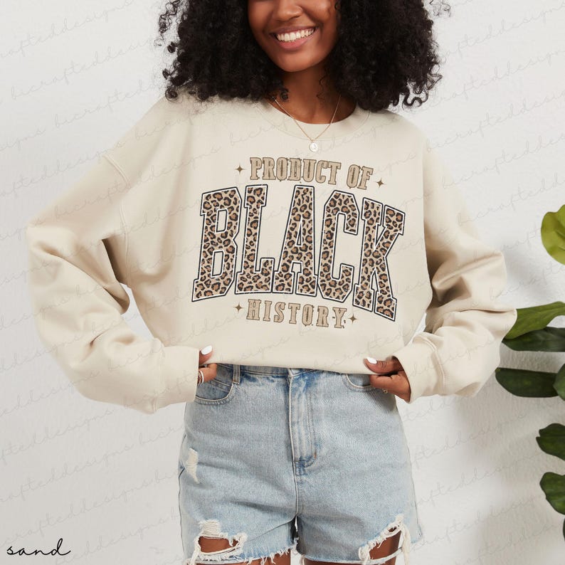 May include: Sand-colored sweatshirt with the text "PRODUCT OF BLACK HISTORY" in a bold, leopard print design. The sweatshirt has long sleeves and a crew neck. The overall style is casual and comfortable.
