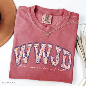 May include: A crimson Comfort Colors t-shirt featuring the text "WWJD" in a floral design. The phrase "He would love first" is below. A gold pendant necklace is draped across the shirt.