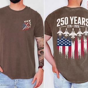 May include: A brown short-sleeve t-shirt featuring a small graphic on the front left chest of fighter jets and a distressed American flag. The back displays the text 250 YEARS 1776-2026 above five fighter jets and a large distressed American flag.