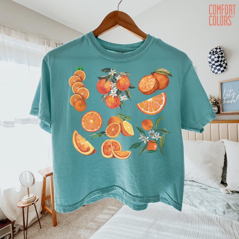 May include: A teal Comfort Colors t-shirt featuring a watercolor illustration of oranges and orange slices. The design includes whole oranges, cut segments, and orange blossoms. The shirt is hanging on a wooden hanger.