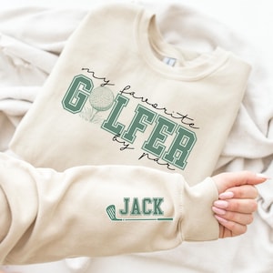 K&ouml;nnte beinhalten: Ein beiger Pullover mit einer gr&uuml;nen und wei&szlig;en Grafik, auf der "my favorite golfer by par" steht, und einem gr&uuml;nen Golfschl&auml;ger mit dem Namen "JACK" auf dem &Auml;rmel.