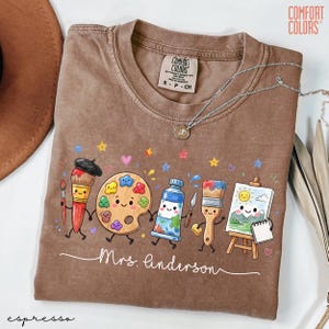 May include: A brown Comfort Colors t-shirt featuring a graphic of anthropomorphic art supplies including a paintbrush, palette, paint tube, and easel with a sun and mountain drawing. The text Mrs. Anderson is written in white cursive script.