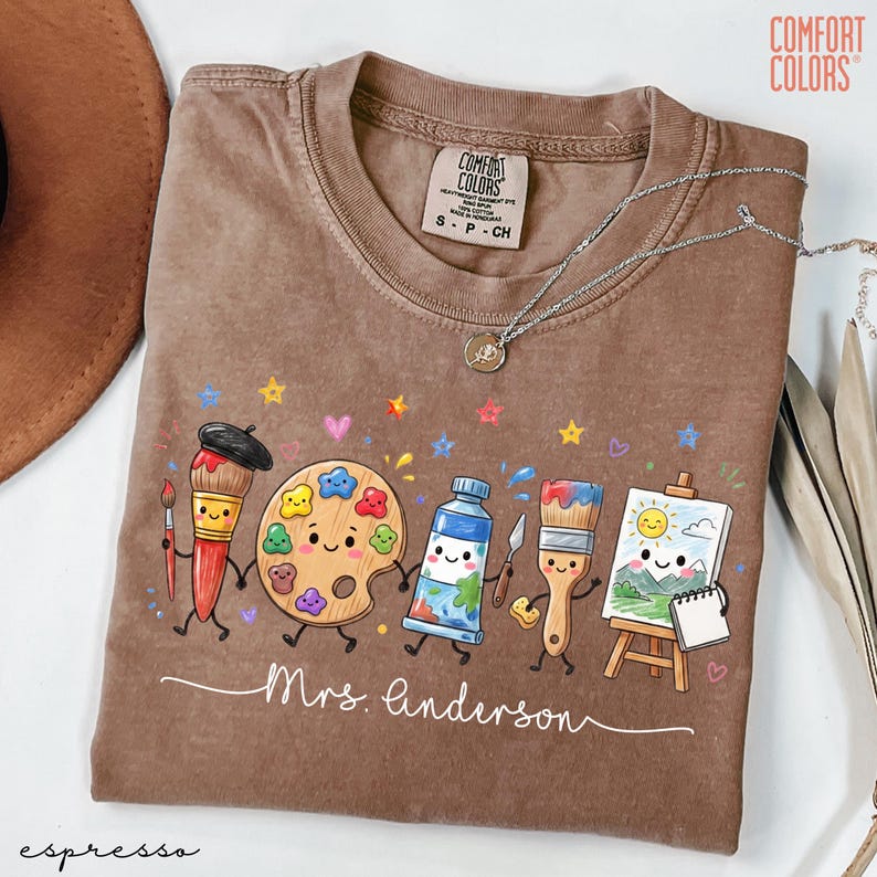 May include: A brown Comfort Colors t-shirt featuring a graphic of anthropomorphic art supplies including a paintbrush, palette, paint tube, and easel with a sun and mountain drawing. The text Mrs. Anderson is written in white cursive script.