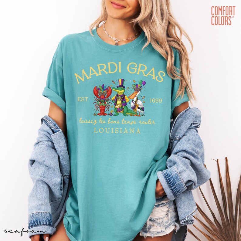 May include: Seafoam green t-shirt with a Mardi Gras design. The design includes the text "MARDI GRAS" above an illustration of a crawfish, alligator, and pelican wearing festive attire. The text "laissez les bons temps rouler LOUISIANA" is below the illustration.