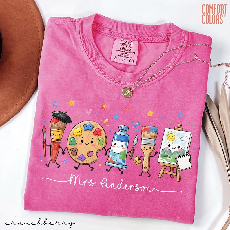 May include: A bright pink Comfort Colors t-shirt featuring a graphic of five anthropomorphic art supplies holding hands, including a paintbrush, palette, paint tube, brush, and easel with a sun painting. The text Mrs. Anderson is written below.
