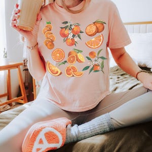 May include: A light pink t-shirt featuring a vibrant orange fruit design. The design includes whole oranges, orange slices, and orange blossoms. The person is wearing light-colored leggings and orange-colored slippers.