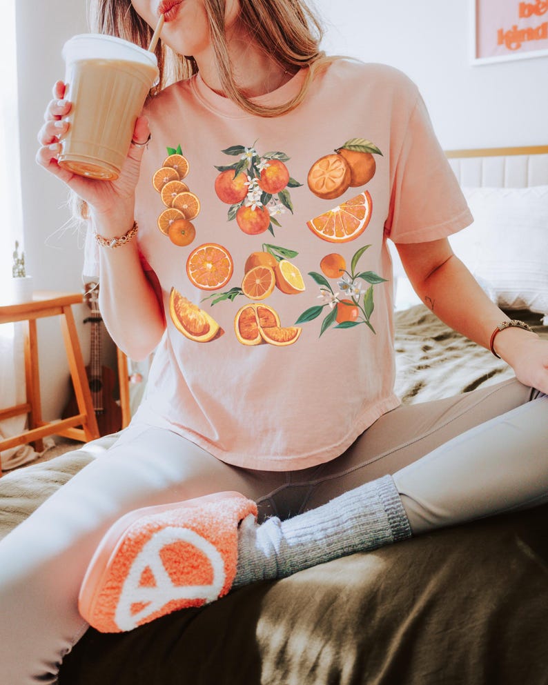 May include: A light pink t-shirt featuring a vibrant orange fruit design. The design includes whole oranges, orange slices, and orange blossoms. The person is wearing light-colored leggings and orange-colored slippers.