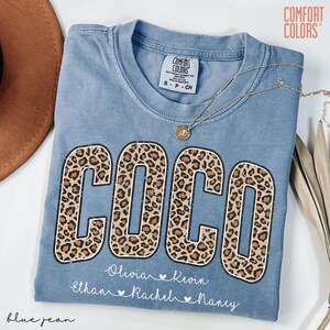 May include: Blue jean Comfort Colors t-shirt with the name "COCO" in large leopard print letters. Below the name are the names Olivia, Kevin, Ethan, Rachel, and Nancy. A gold necklace with a pendant is draped across the shirt.