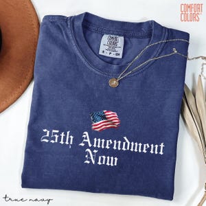 May include: A true navy blue t-shirt with the text "25th Amendment Now" and an American flag graphic. The shirt is made of Comfort Colors material and has a small tag at the neckline. A gold necklace is draped across the shirt.