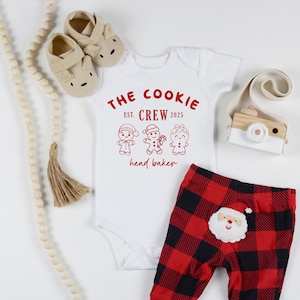 May include: A white baby onesie with the text "THE COOKIE CREW EST. 2025 head baker" in red, paired with red and black plaid pants featuring a Santa Claus applique. Includes beige baby shoes and a wooden toy camera.
