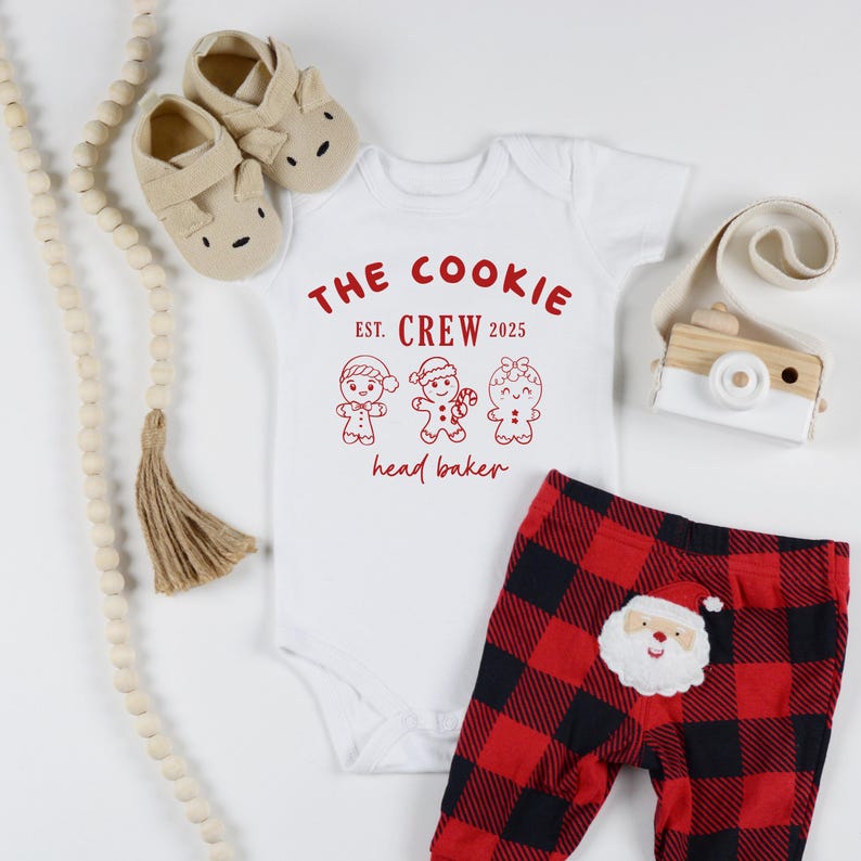 May include: A white baby onesie with the text "THE COOKIE CREW EST. 2025 head baker" in red, paired with red and black plaid pants featuring a Santa Claus applique. Includes beige baby shoes and a wooden toy camera.