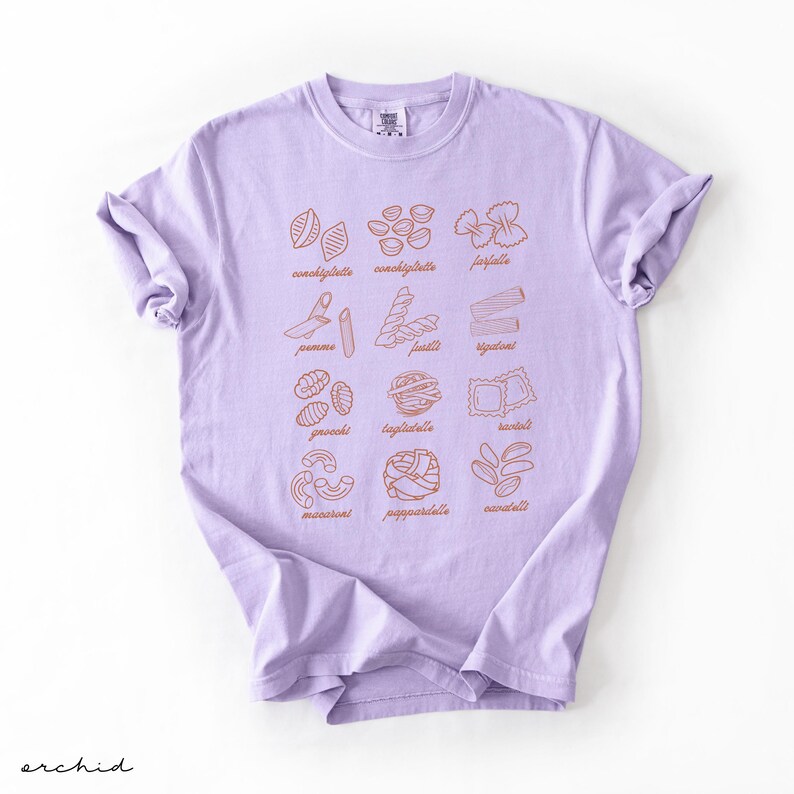 Op de afbeelding: Een lavendelkleurig T-shirt met een afbeelding van verschillende soorten pasta met hun namen onder elke afbeelding. De pastasoorten zijn:  conchiglie,  conchigliette,  farfalle,  penne,  fusilli,  spaghetti,  gnocchi,  tagliatelle,  tortellini,  macaroni,  pappardelle  en  cannelloni.