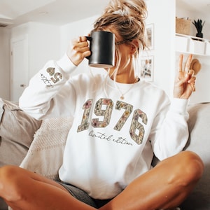 May include: White sweatshirt with the year "1976" in a floral design, with the words "limited edition" below. The left sleeve has the text "Chapter 50". A black mug is held up to the face.