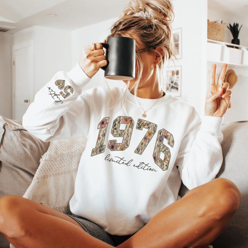 May include: White sweatshirt with the year "1976" in a floral design, with the words "limited edition" below. The left sleeve has the text "Chapter 50". A black mug is held up to the face.