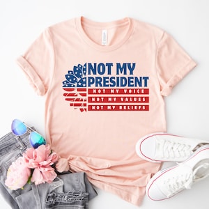 May include: A pink t-shirt with a sunflower design featuring the American flag colors. The text on the shirt reads "Not My President, Not My Voice, Not My Values, Not My Beliefs".