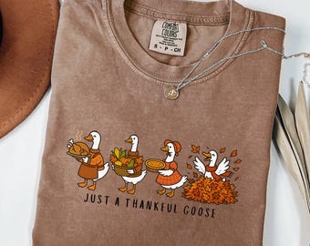 May include: Brown Comfort Colors t-shirt featuring a Thanksgiving-themed graphic. The design shows four geese with a turkey, basket of vegetables, pie, and leaves. The text reads "JUST A THANKFUL GOOSE". A silver necklace is draped across the shirt.