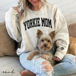 May include: A sand-colored sweatshirt with the text "YORKIE MOM" in black block letters. The sleeve has the name "Scout Daisy" and a paw print. A small Yorkshire Terrier dog is in the image.