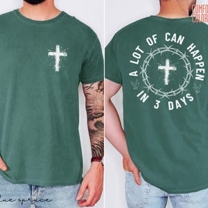 May include: A blue spruce t-shirt with a white cross graphic on the front and back. The back of the shirt features the text "A LOT OF CAN HAPPEN IN 3 DAYS" surrounding a cross and a crown of thorns.