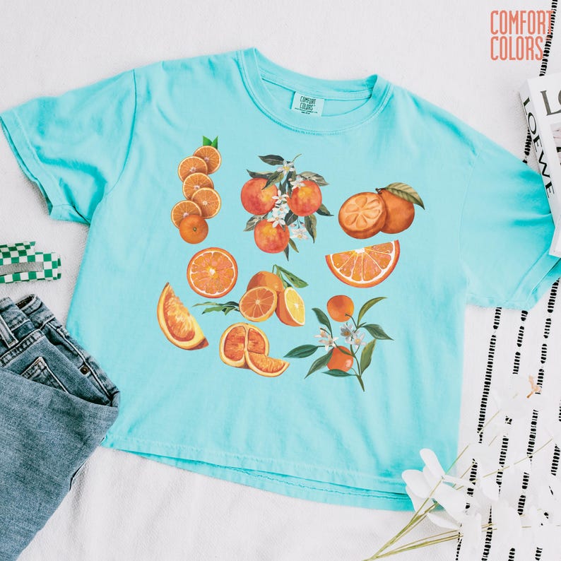 May include: A light blue t-shirt featuring a vibrant orange fruit design. The graphic includes whole oranges, slices, and orange blossoms. The shirt is made of a soft material and has the words "COMFORT COLORS" printed on the upper right.