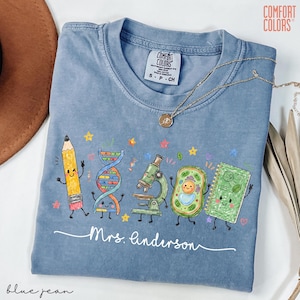 May include: A blue Comfort Colors t-shirt featuring a graphic of a cartoon pencil, DNA strand, microscope, cell, and notebook with faces. The text "Mrs. Anderson" is written in white script below the illustrations. A gold necklace rests on the collar.