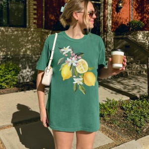 May include: A green t-shirt featuring a watercolor illustration of lemons, white blossoms, and green leaves. The shirt is a short-sleeved, casual style. The lemons are yellow, and the blossoms are white and pink.