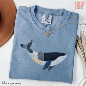 May include: A blue jean Comfort Colors t-shirt featuring an appliqued whale design. The whale is made of various shades of blue and gray fabric. A gold necklace with a pendant rests on the shirt. The text "blue jean" is at the bottom.
