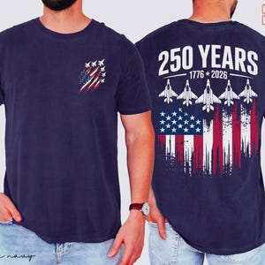 May include: A navy blue t-shirt featuring a small graphic on the left chest of a distressed American flag with fighter jets. The back displays the text 250 YEARS 1776-2026 above five fighter jets flying over a large distressed American flag.
