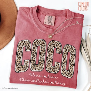 May include: Crimson-colored t-shirt with the name "COCO" in large leopard print letters. Below the name are smaller names: Olivia, Kevin, Ethan, Rachel, and Nancy. The shirt has a crew neck and the Comfort Colors brand label.