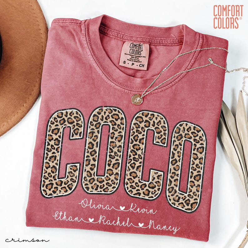 May include: Crimson-colored t-shirt with the name "COCO" in large leopard print letters. Below the name are smaller names: Olivia, Kevin, Ethan, Rachel, and Nancy. The shirt has a crew neck and the Comfort Colors brand label.