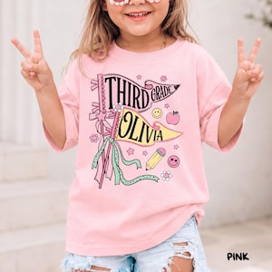 May include: A light pink t-shirt featuring a graphic print of two pennant flags with the text "THIRD GRADE" and "OLIVIA". The design includes a yellow pencil, a pink apple, two smiley faces, and small flowers, all accented with ribbons.