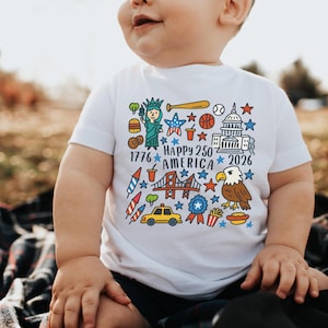 K&ouml;nnte beinhalten: Wei&szlig;es T-Shirt mit einem farbenfrohen Grafikdesign, das den Text "Happy 250 America 2026" und Illustrationen der Freiheitsstatue, eines Baseballschl&auml;gers und eines Adlers enth&auml;lt. Das Design zeigt auch Sterne und andere patriotische Symbole. F&uuml;r Kinder.
