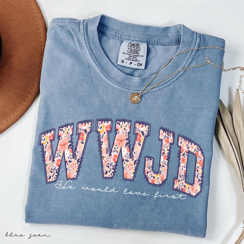 May include: Blue Comfort Colors t-shirt with the letters WWJD in a floral design. The text "He would love first" is below the letters. A gold necklace with a pendant is on the shirt.