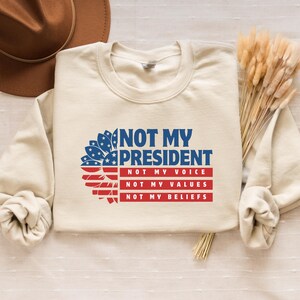 May include: A beige sweatshirt with a sunflower design featuring the American flag colors. The text "NOT MY PRESIDENT" is printed in blue, with the words "NOT MY VOICE", "NOT MY VALUES", and "NOT MY BELIEFS" printed in red below.