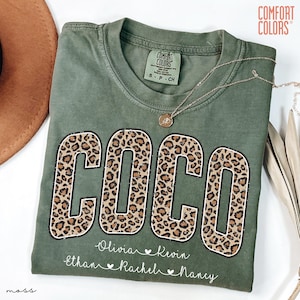 May include: Olive green Comfort Colors t-shirt with the name "COCO" in large leopard print letters. Below the name are smaller names: Olivia, Kevin, Ethan, Rachel, and Nancy. A gold necklace rests on the shirt.