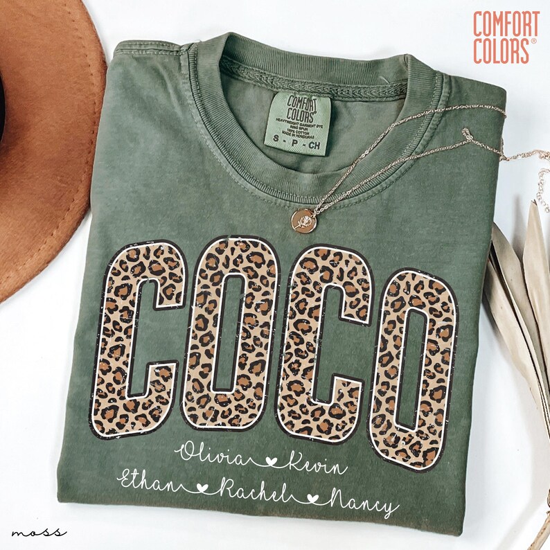 May include: Olive green Comfort Colors t-shirt with the name "COCO" in large leopard print letters. Below the name are smaller names: Olivia, Kevin, Ethan, Rachel, and Nancy. A gold necklace rests on the shirt.