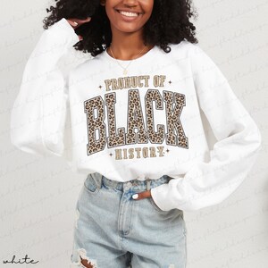 May include: White sweatshirt with long sleeves featuring the text "PRODUCT OF BLACK HISTORY" in a bold, leopard print design. The sweatshirt has a crew neck and a relaxed fit, suitable for casual wear.