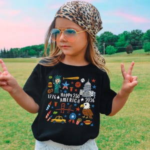 K&ouml;nnte beinhalten: Ein schwarzes T-Shirt mit einer farbenfrohen Grafik, die den 250. Jahrestag Amerikas feiert. Das Design umfasst die Freiheitsstatue, einen Baseballschl&auml;ger, einen Basketball, das Kapitol, einen Adler und den Text "1776 HAPPY 250 AMERICA 2026". Die Person tr&auml;gt eine Pilotenbrille und ein Leoparden-Kopftuch.