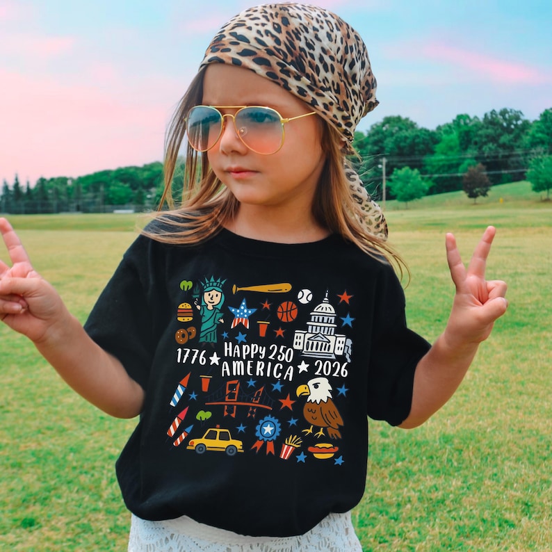 K&ouml;nnte beinhalten: Ein schwarzes T-Shirt mit einer farbenfrohen Grafik, die den 250. Jahrestag Amerikas feiert. Das Design umfasst die Freiheitsstatue, einen Baseballschl&auml;ger, einen Basketball, das Kapitol, einen Adler und den Text "1776 HAPPY 250 AMERICA 2026". Die Person tr&auml;gt eine Pilotenbrille und ein Leoparden-Kopftuch.
