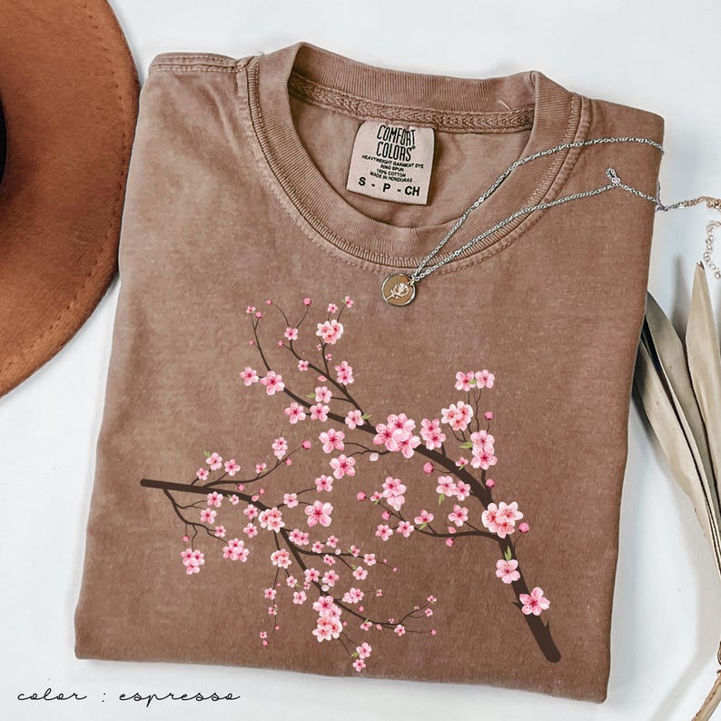 May include: A brown t-shirt with a pink cherry blossom branch design. The shirt has a distressed look and the text "COMFORT COLORS" is visible on the tag. The color of the shirt is "espresso".